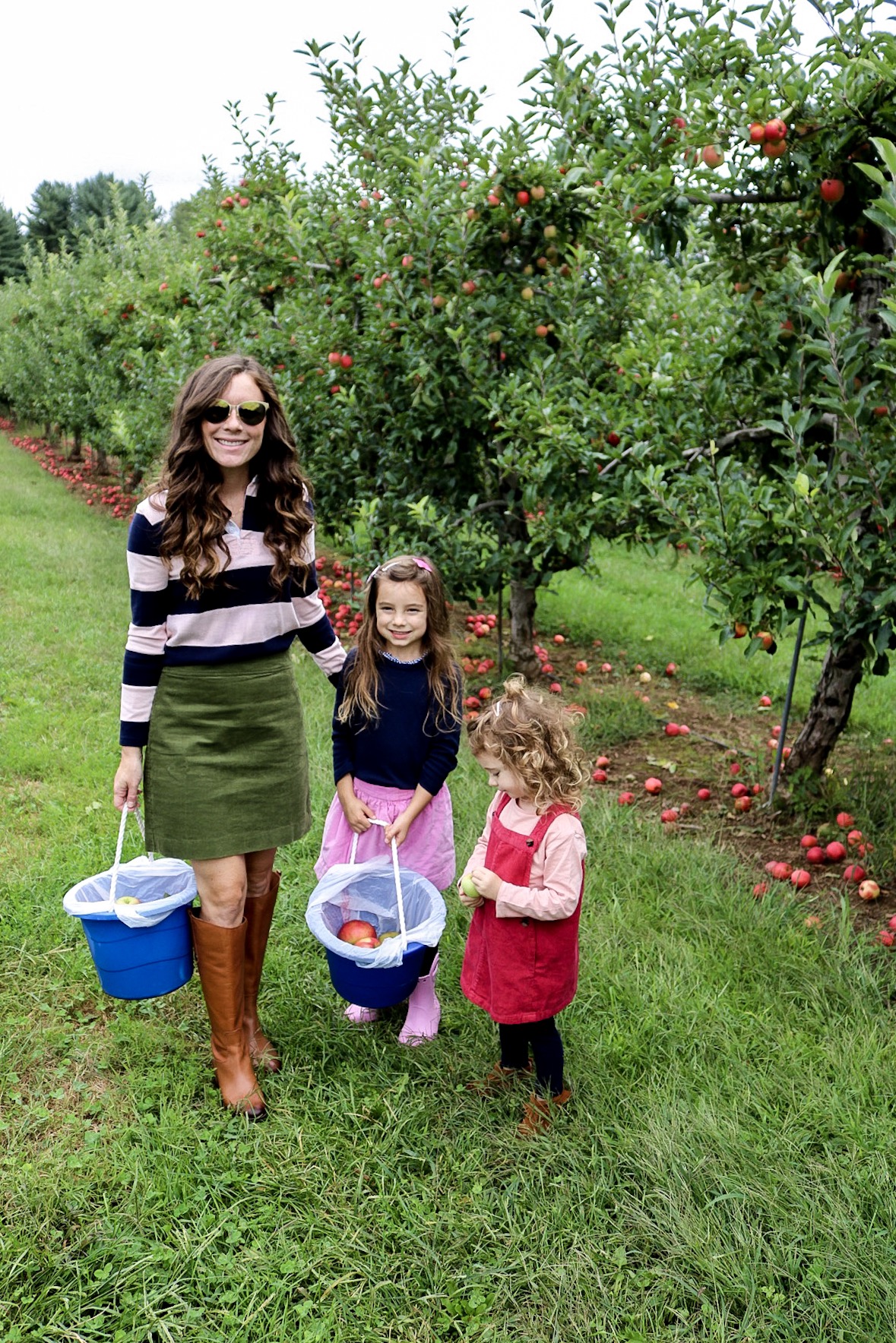 Woman in boots and corduroy skirt apple picking with little girls wearing dresses