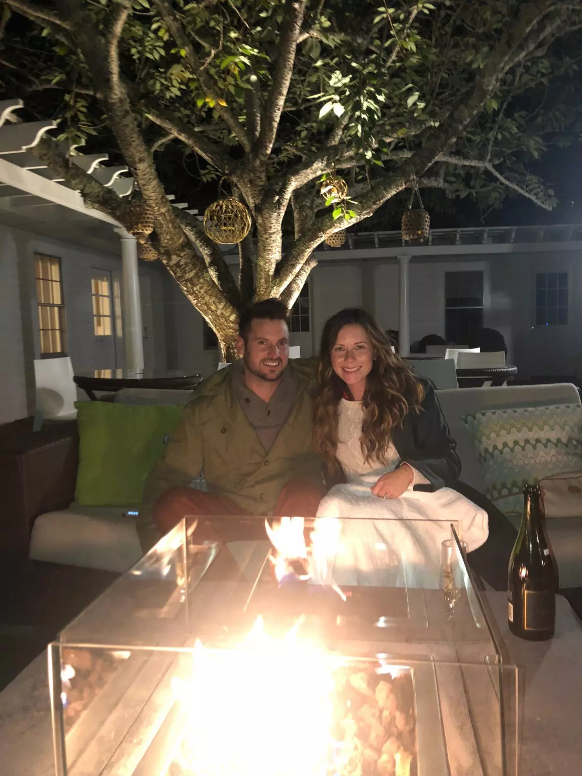 Man and Woman sitting under a blanket on a bench by a fire pit in a courtyard