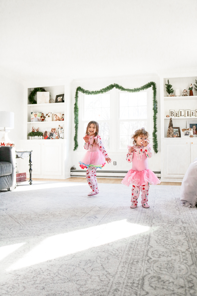Little girls wearing pink pajamas and pink tutus dancing and laughing in the middle of a large very white and bright family room decorated for Christmas